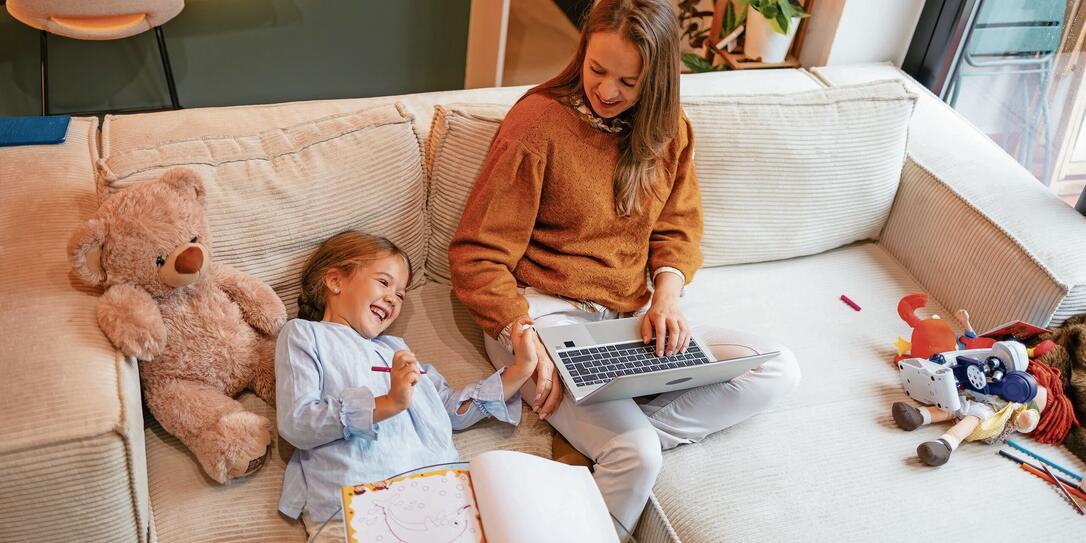 Mother and daughter are smiling while working and playing together on a sofa.