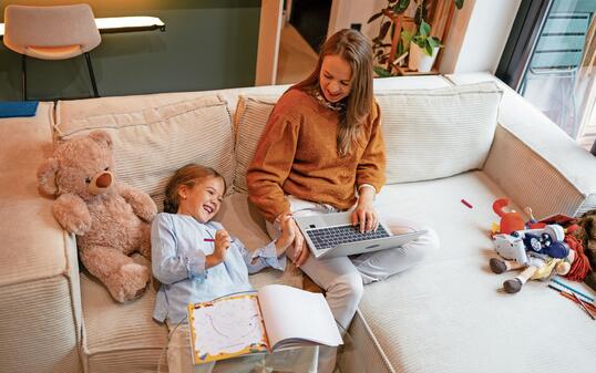 Mother and daughter are smiling while working and playing together on a sofa.