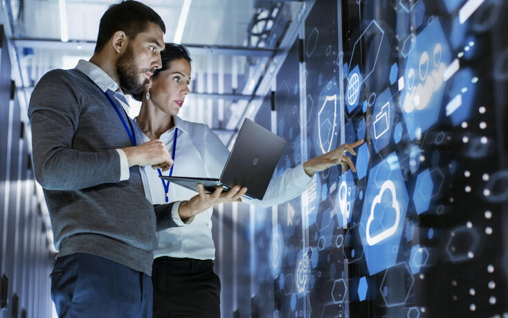 Male IT Specialist Holds Laptop and Discusses Work with Female Server Technician. They're Standing in Data Center, Rack Server Cabinet with Cloud Server Icon and Visualization.