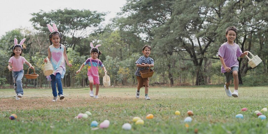 Happy group of diverse cute little children hunting Easter eggs, wearing bunny ears. kids holding basket, running to collect eggs on grass while playing outdoors at park