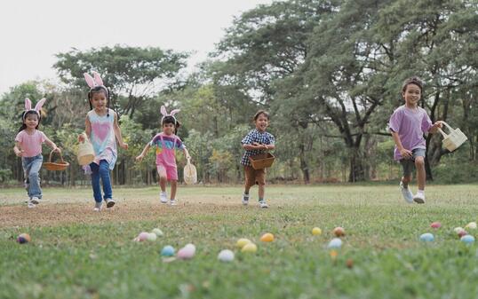 Happy group of diverse cute little children hunting Easter eggs, wearing bunny ears. kids holding basket, running to collect eggs on grass while playing outdoors at park