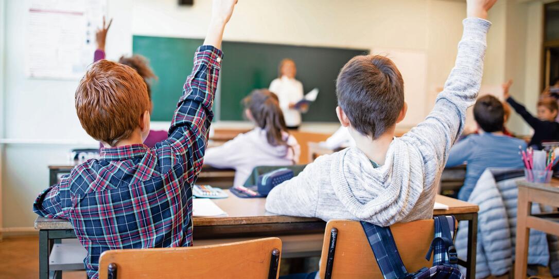 Schoolchildren at classroom with raised hands answering teacher's question.