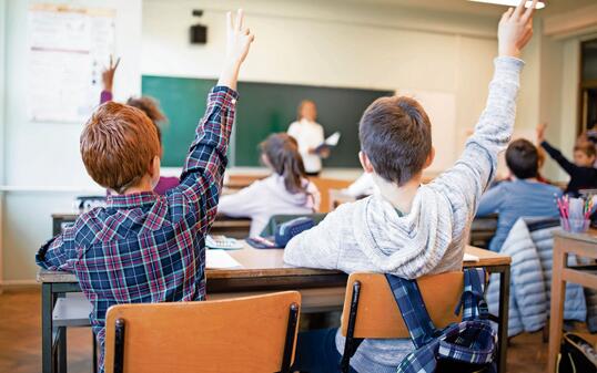 Schoolchildren at classroom with raised hands answering teacher's question.