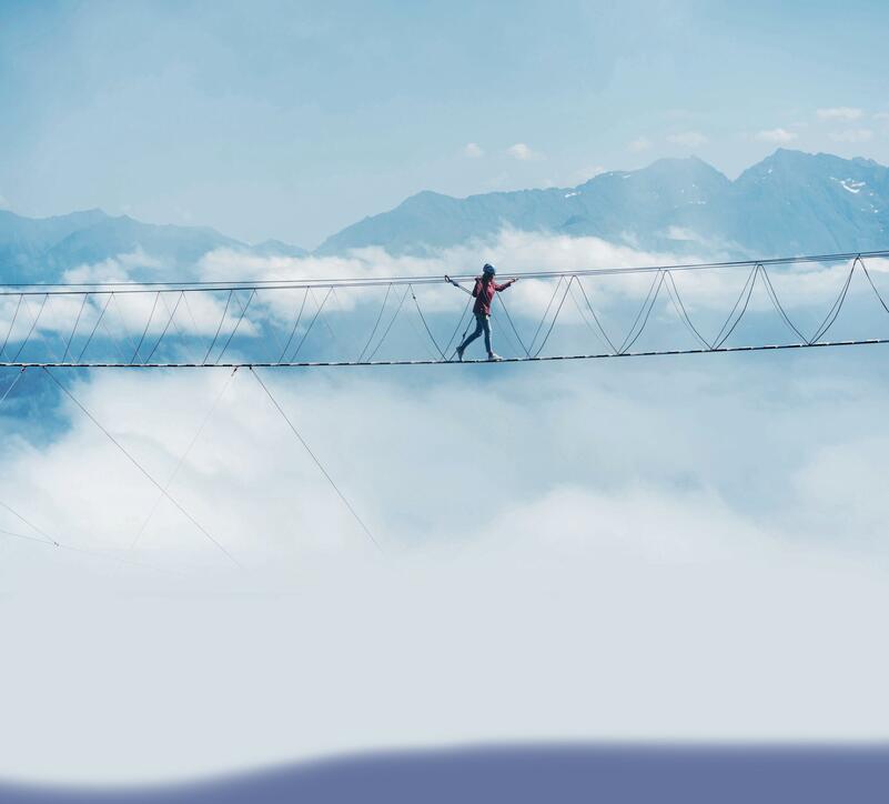 A person walks on a suspended rope bridge in the clouds.