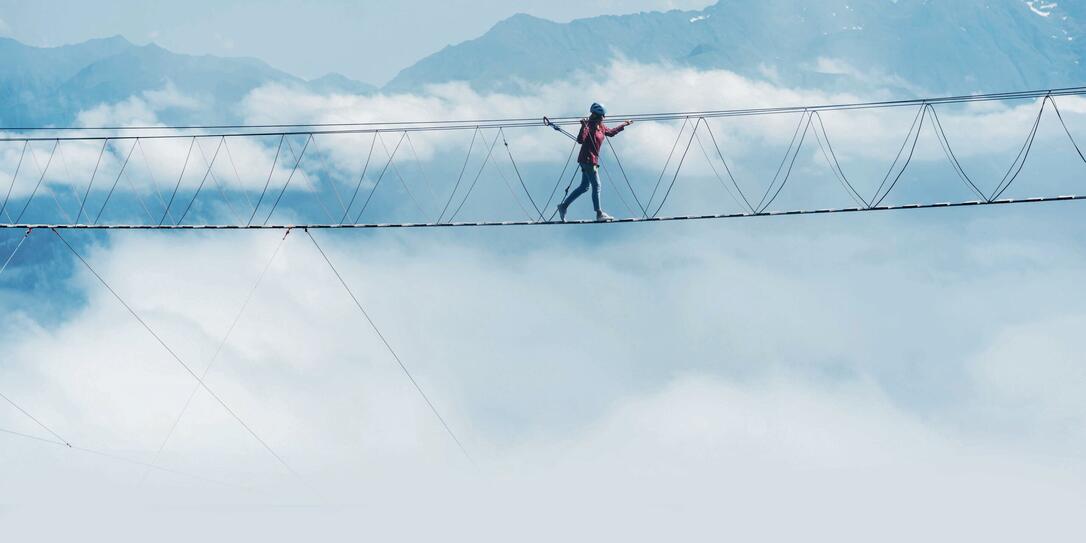 A person walks on a suspended rope bridge in the clouds.