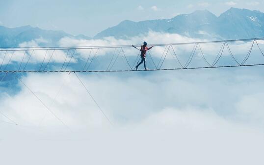 A person walks on a suspended rope bridge in the clouds.