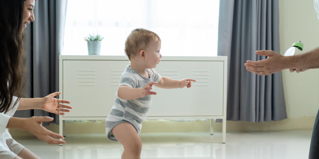 Caucasian young cute baby boy child learn to walk with parent in house. Happy family mother support little toddler son taking first step walk on floor to father to develop skill in living room at home Caucasian young cute baby boy child learn to walk with parent in house. Happy family mother support little toddler son taking first step walk on floor to father to develop skill in living room at home