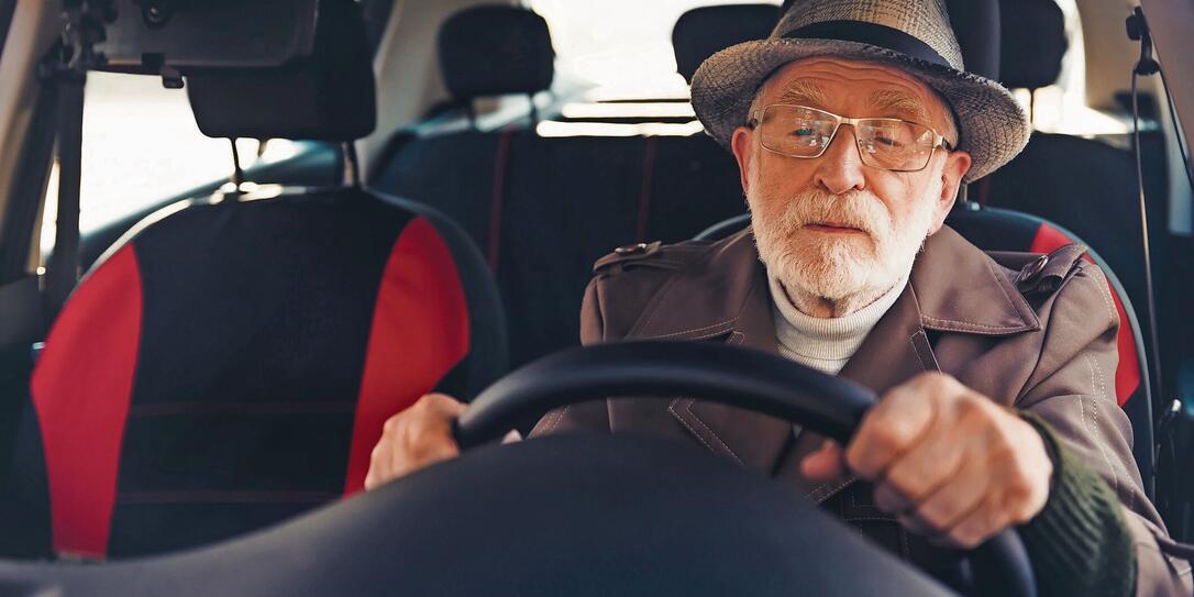 Elderly man wearing a hat driving a vehicle on a sunny day displaying focus and confidence on an urban road