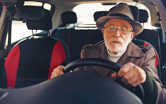 Elderly man wearing a hat driving a vehicle on a sunny day displaying focus and confidence on an urban road