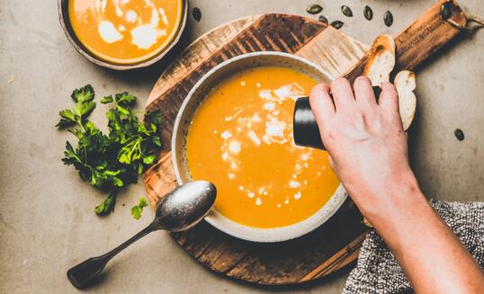 Mans hand adding pepper to pumpkin cream soup with seeds