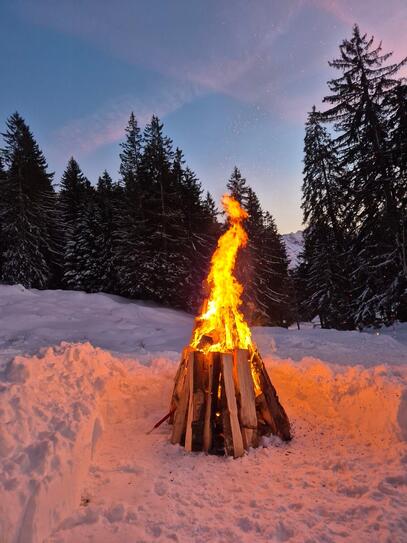A bonfire burning in a snowy landscape at dusk. The flames cast a warm glow on the snow, with tall evergreen trees and a mountain in the background under a fading blue and pink sky.