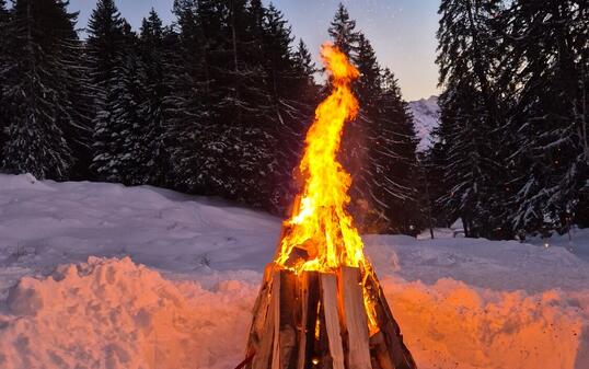 A bonfire burning in a snowy landscape at dusk. The flames cast a warm glow on the snow, with tall evergreen trees and a mountain in the background under a fading blue and pink sky.