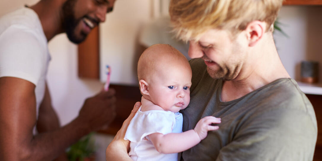Loving Male Same Sex Couple Cuddling Baby Daughter In Bathroom At Home Together