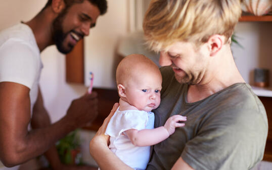 Loving Male Same Sex Couple Cuddling Baby Daughter In Bathroom At Home Together