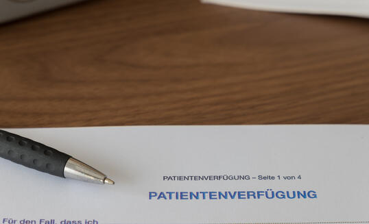 Patientenverfügung Seite 1 von 4 means living will page 1 of 4, the blue headline living will, with the german translation "In Case that I...", a pen and books lie on the table