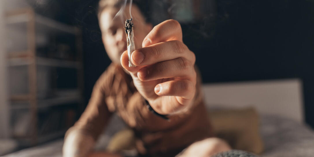 Man offering a hand rolled cannabis cigarette while sitting at the bed