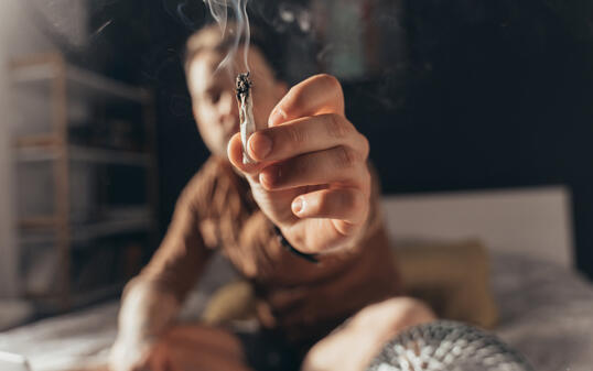 Man offering a hand rolled cannabis cigarette while sitting at the bed