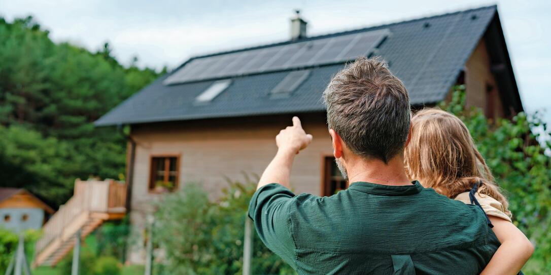 Rear view of dad holding her little girl in arms and showing at their house with installed solar panels. Alternative energy, saving resources and sustainable lifestyle concept. Rear view of dad holding her little girl in arms and showing at their house with installed solar panels. Alternative energy, saving resources and sustainable lifestyle concept.