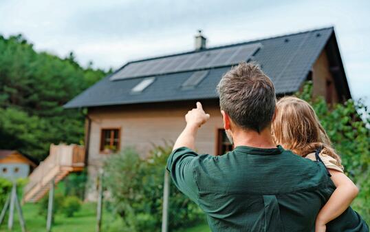 Rear view of dad holding her little girl in arms and showing at their house with installed solar panels. Alternative energy, saving resources and sustainable lifestyle concept.