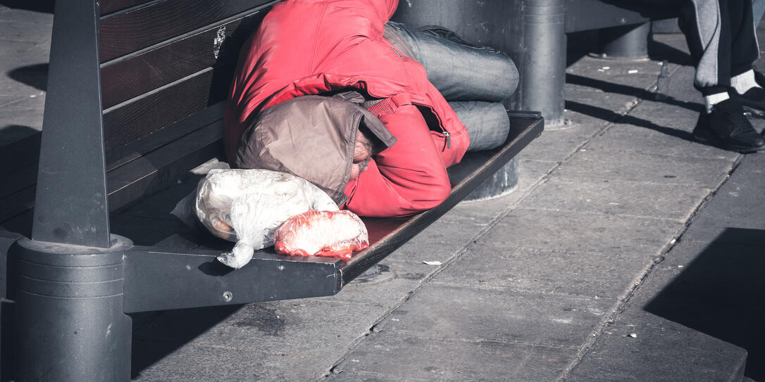 Poor homeless man sleeps on the wooden bench at the street in the city in red winter jacket, social documentary concept