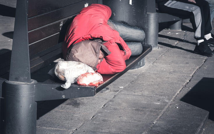 Poor homeless man sleeps on the wooden bench at the street in the city in red winter jacket, social documentary concept