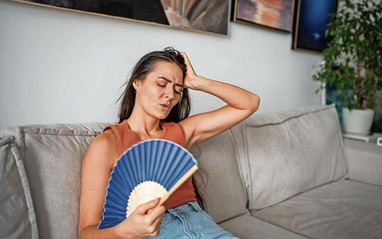Overheated woman blowing with paper fan, sitting on couch at home, trying to cool down at home