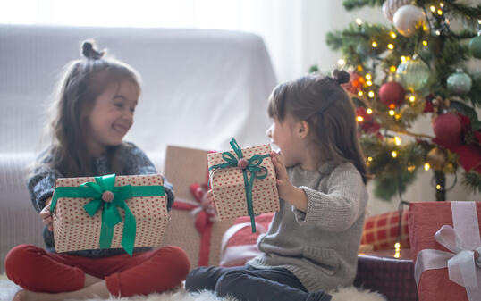 Pretty little girls are holding a gift boxes and smiling at home on light background