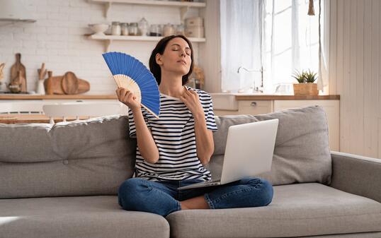 Young woman suffers from heatstroke flat without air-conditioner waving fan sitting on couch at home