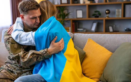 Couple sitting on the sofa and embracing. Soldier in uniform is at home with his wife. Flag of Ukraine.