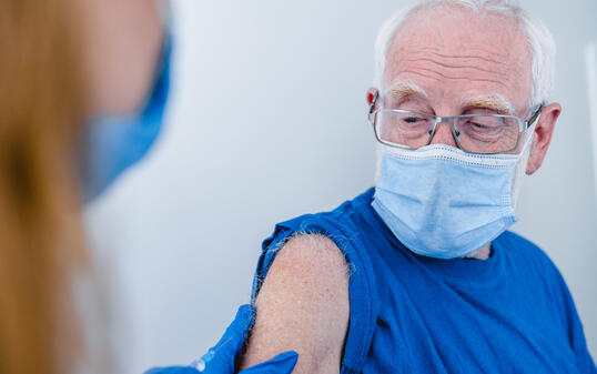 Close up photo of senior man`s arm with doctor`s hands making injection. Elderly patient wearing mask to stop coronavirus spread