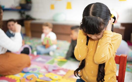 Preschool girl covers her ears and closes her eyes while other kindergarten children play loudly behind her. Concept of noise sensitivity, sensory overload, and emotional response in early childhood