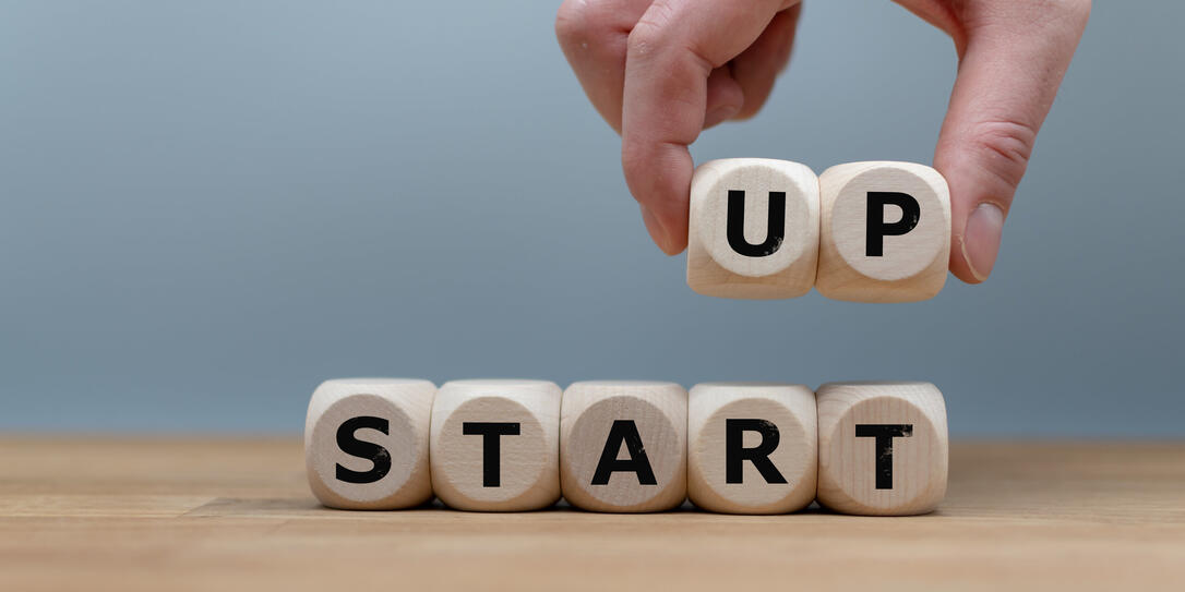 Cubes form the words "START UP" while to fingers lift the letters "UP" in the air. Cubes are on a wooden table in-front a grey background.