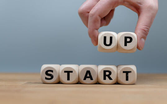 Cubes form the words "START UP" while to fingers lift the letters "UP" in the air. Cubes are on a wooden table in-front a grey background.