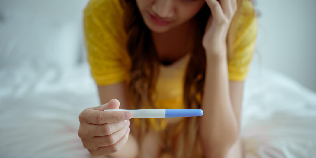 Young asian young woman feeling shock after look at positive Pregnancy test. Disappointed unplanned pregnancy girl sit on bed with upset and sad face for problem don't want to pregnant.