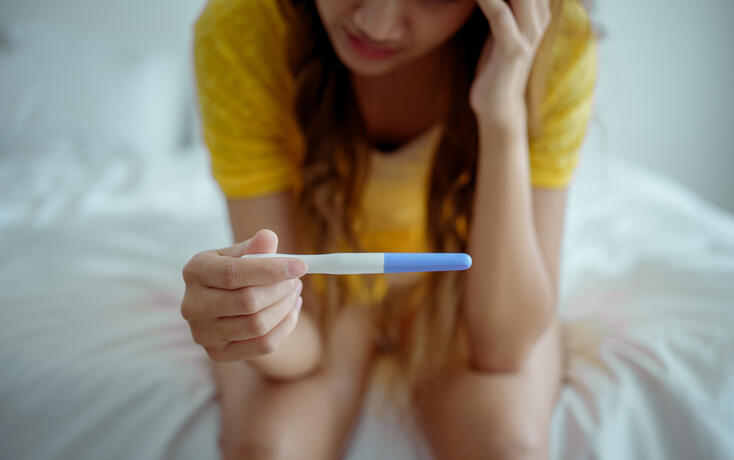 Young asian young woman feeling shock after look at positive Pregnancy test. Disappointed unplanned pregnancy girl sit on bed with upset and sad face for problem don't want to pregnant.