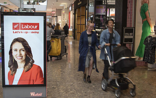 Menschen gehen in einem Einkaufszentrum an einer elektronischen Plakatwand vorbei, die ein Foto der neuseeländischen Premierministerin Jacinda Ardern zeigt. Neuseeländer gehen bei den Parlamentswahlen am Samstag, den 17.10.2020, an die Urnen. Foto: Mark Baker/AP/dpa