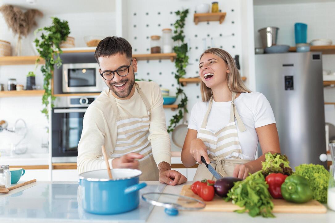 Beautiful young couple is feeding each other and smiling while cooking in kitchen at home. Happy sporty couple is preparing healthy food on light kitchen. Healthy food concept.