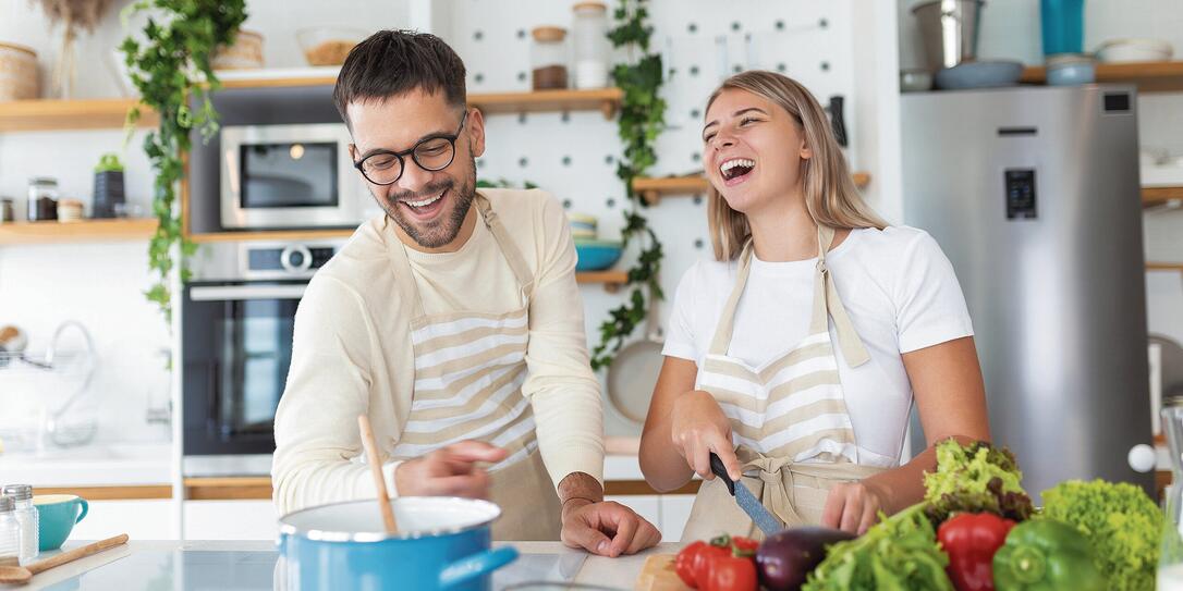 Beautiful young couple is feeding each other and smiling while cooking in kitchen at home. Happy sporty couple is preparing healthy food on light kitchen. Healthy food concept.