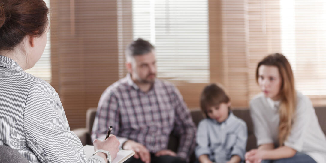 Rear view of female psychologist helping young family with a kid to solve child development problems. Family sitting on a sofa in the blurred background
