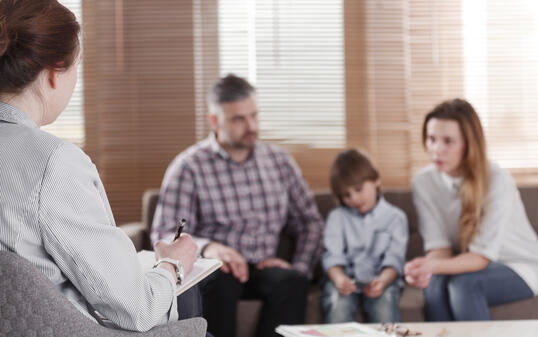 Rear view of female psychologist helping young family with a kid to solve child development problems. Family sitting on a sofa in the blurred background