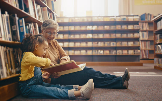 Library, learning and grandmother with grandchild on floor with book for reading, story and fun together. Education, child development and grandma with girl, storytelling and teaching while bonding