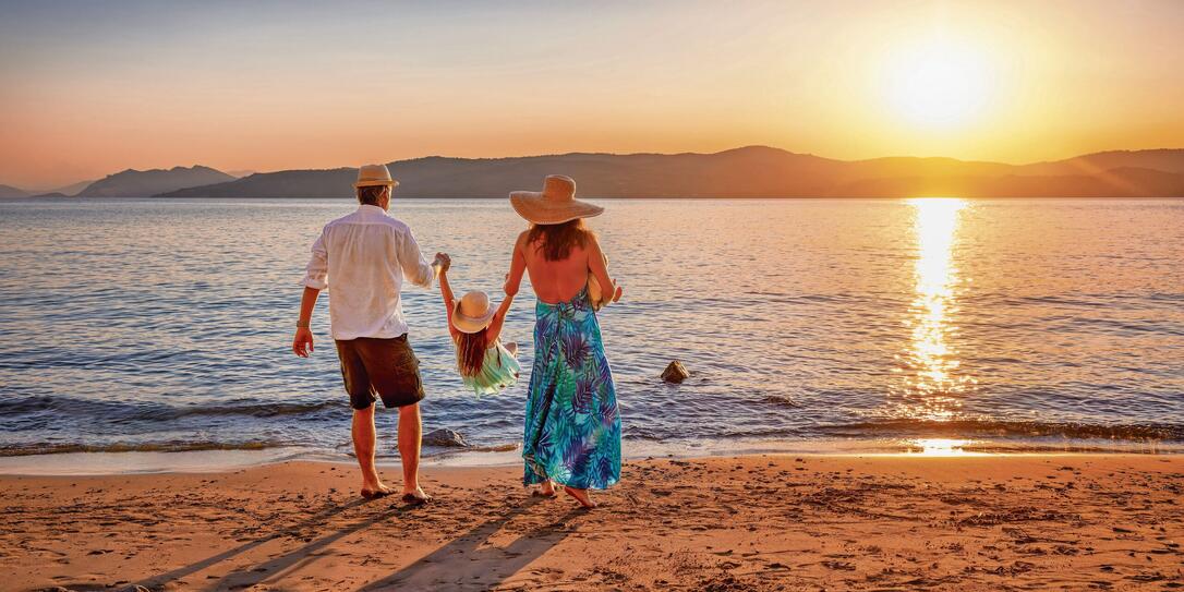 A family on holidays stands on a beach in the mediterranean sea and enjoys the summer sunset time