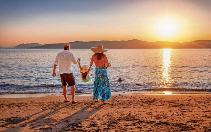 A family on holidays stands on a beach in the mediterranean sea and enjoys the summer sunset time