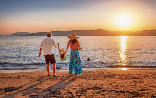 A family on holidays stands on a beach in the mediterranean sea and enjoys the summer sunset time
