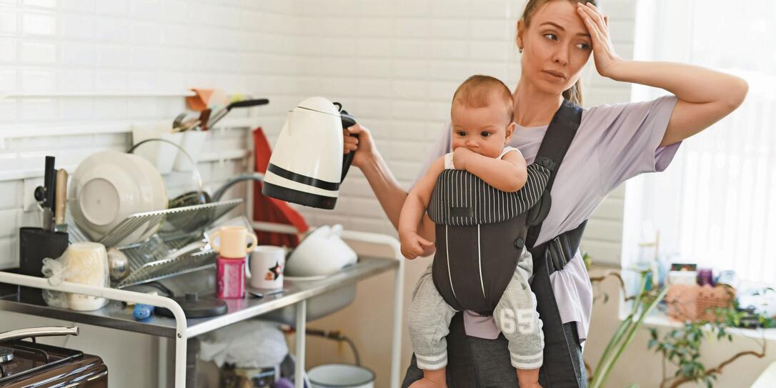 Tired multitasking mother and child in baby carrier. Young girl in the kitchen with a child. The difficulties of motherhood and loneliness
