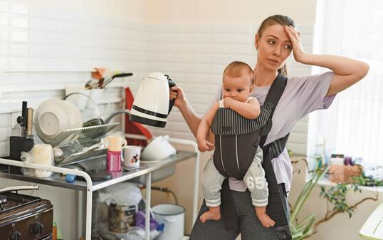 Tired multitasking mother and child in baby carrier. Young girl in the kitchen with a child. The difficulties of motherhood and loneliness