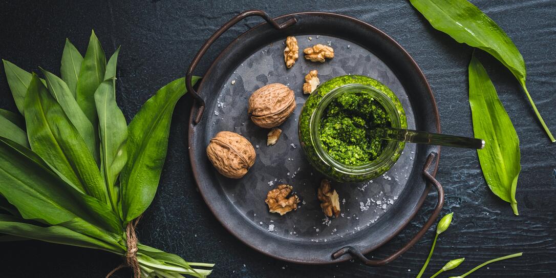 Homemade wild garlic pesto and walnuts on a metal tray, wild garlic leaves and blossoms on black background