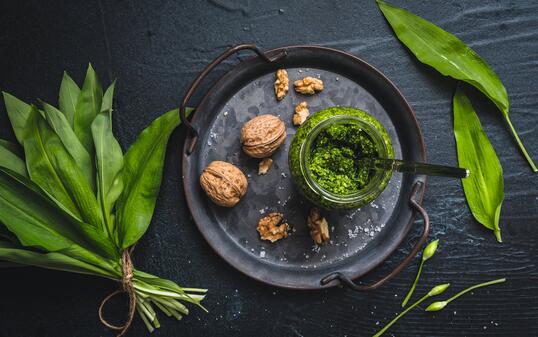 Homemade wild garlic pesto and walnuts on a metal tray, wild garlic leaves and blossoms on black background
