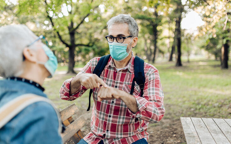 Senior man and his wife with protective face masks resting in the nature and talking on sign language