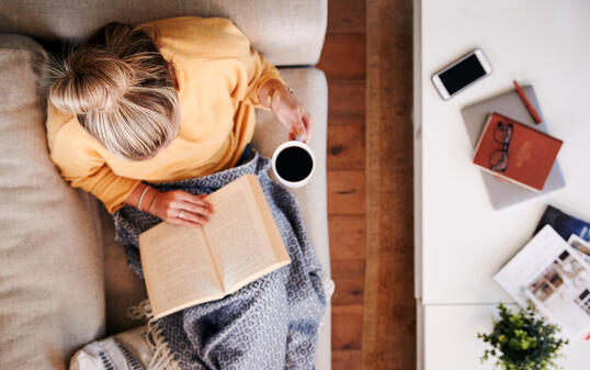 Overhead Shot Looking Down On Woman At Home Lying On Reading Book And Drinking Coffee